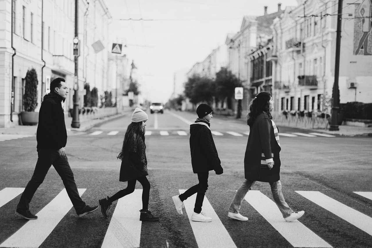 una famiglia che attraversa la strada sulle strisce pedonali ricalcando la foto iconica dei Beatles: la mamma davanti, i due figli - maschio e femmina in età scolare- dietro e ultimo della fila il papà. La foto è in bianco e nero, ambientata in autunno in un contesto urbano